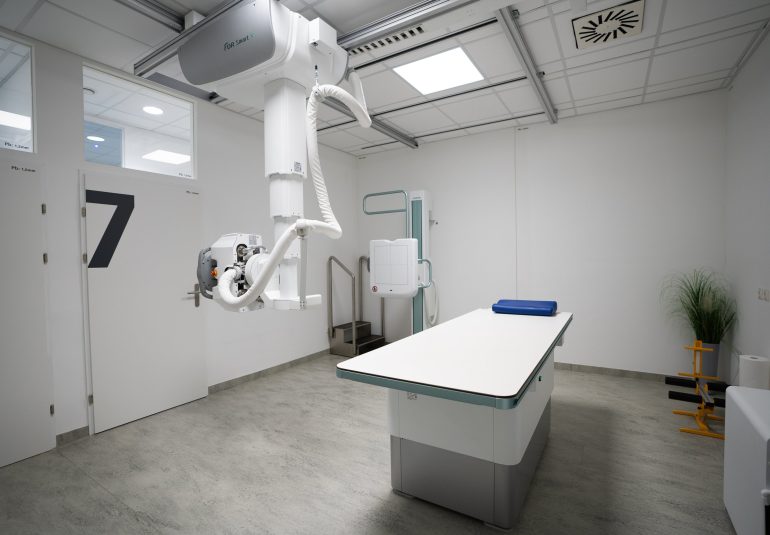 Moderner Röntgenraum in der Radiologiepraxis mit Untersuchungsliege, Deckenstativ-Röntgengerät und Schutzwand.