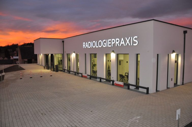 Abendliche Außenansicht der Radiologiepraxis Oberpullendorf mit leuchtendem Praxisschild und wunderschönem Sonnenuntergang