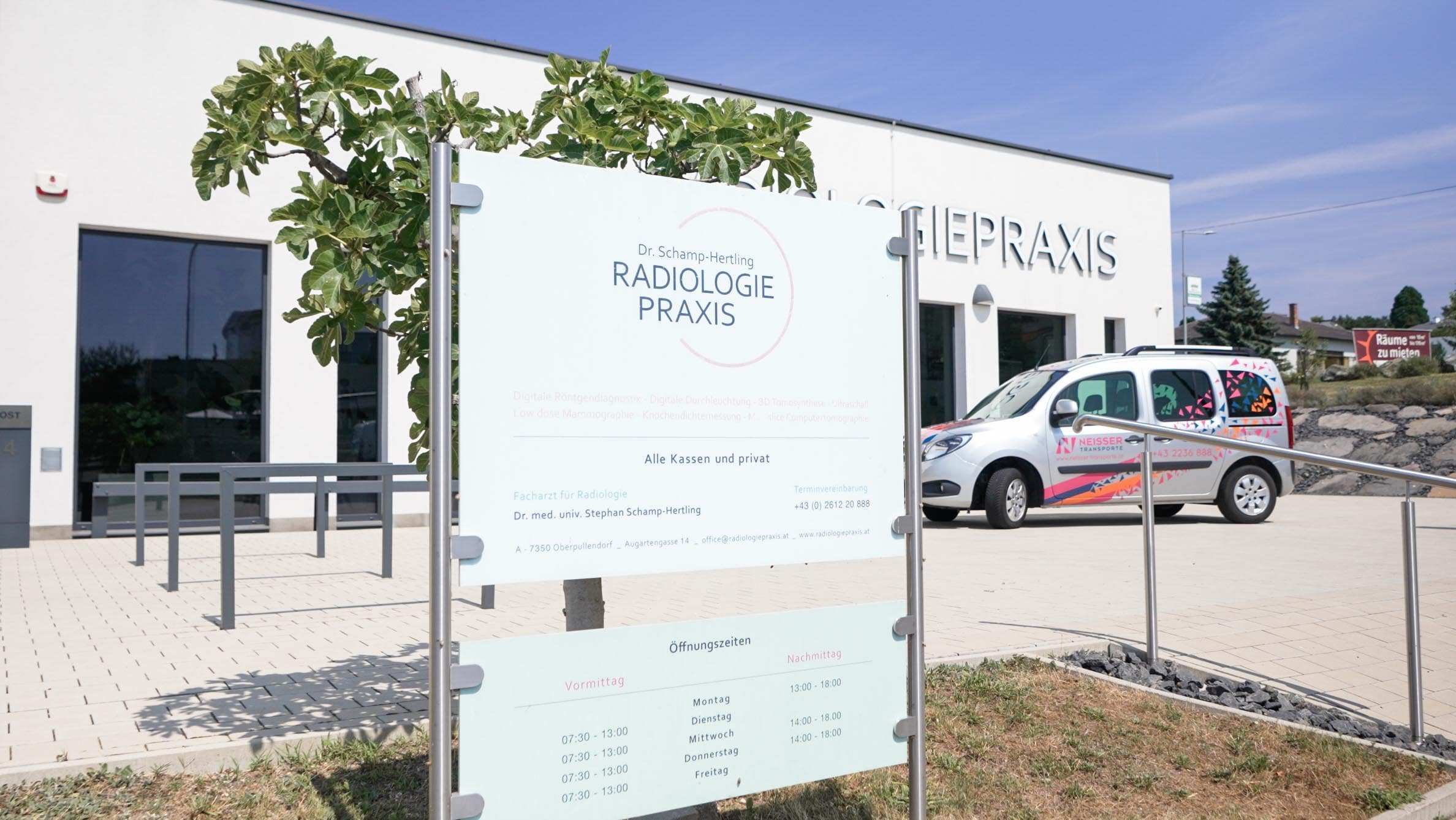 Außenansicht der Radiologiepraxis Dr. Schamp-Hertling in Oberpullendorf mit Praxis-Schild und medizinischem Transportfahrzeug.