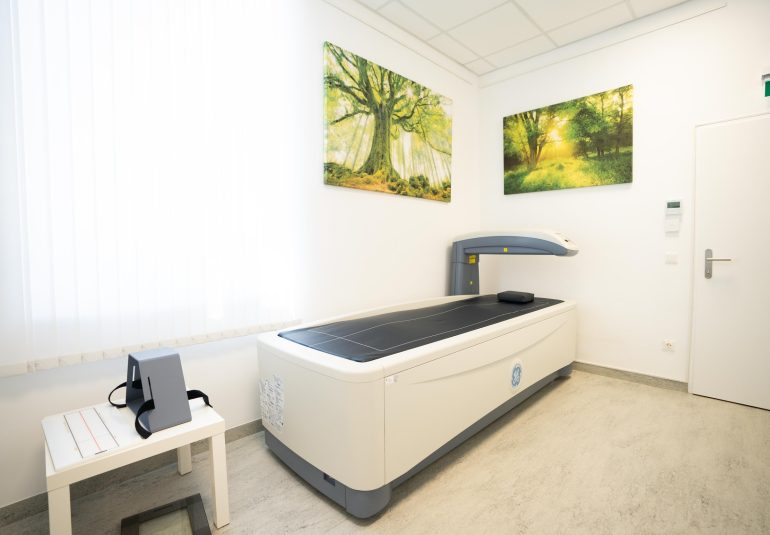 Knochendichtemessgerät in der Radiologiepraxis mit Naturbildern an der Wand und einem kleinen Tisch mit medizinischem Zubehör.