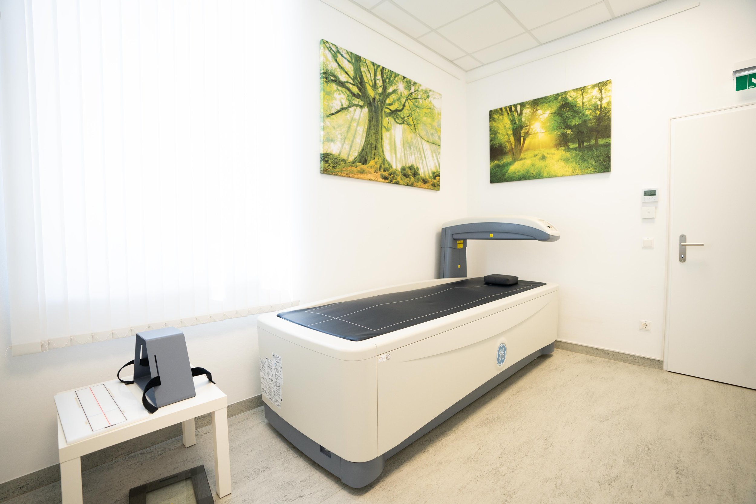 Knochendichtemessgerät in der Radiologiepraxis mit Naturbildern an der Wand und einem kleinen Tisch mit medizinischem Zubehör.