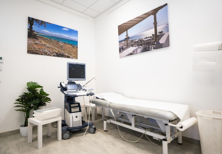 Sonographiegerät in der Radiologiepraxis neben einer Untersuchungsliege, mit Urlaubsbildern an der Wand und medizinischem Zubehör.