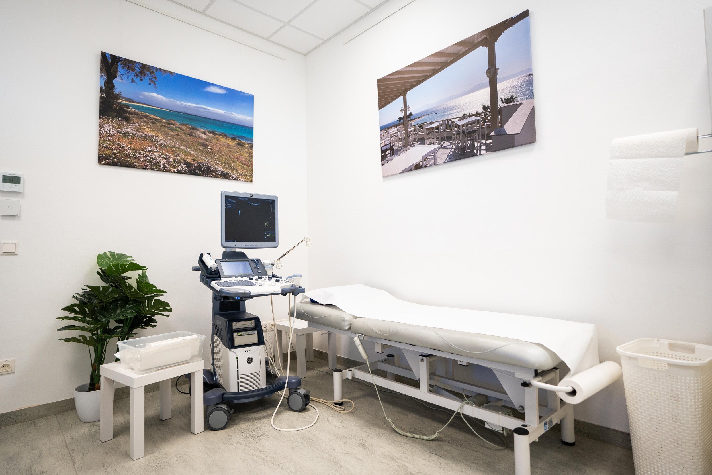 Sonographiegerät in der Radiologiepraxis neben einer Untersuchungsliege, mit Urlaubsbildern an der Wand und medizinischem Zubehör.
